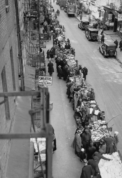 High angle view showing shoppers crowding around the carts of a pushcart market along a sidewalk on the west side of Orchard Street in the Lower East
