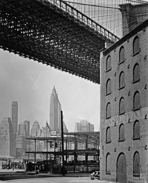 Steel frame building going up under the Brooklyn Bridge next to brick warehouse Manhattan skyline in the distance.