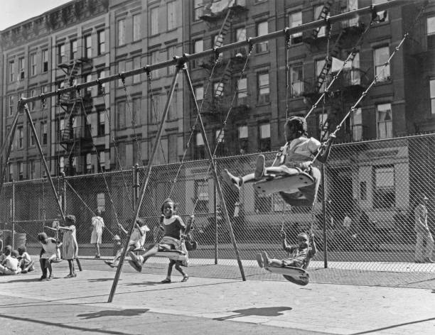 African-American children playing swings at a playground with a chainlink fence in the background and tenement blocks beyond that in Harlem New