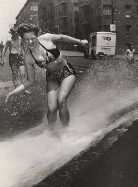 An unidentified woman in a two-piece bathing suit cringes as she stands in a spray of water from a fire hydrant New York 1930s or 1940s.