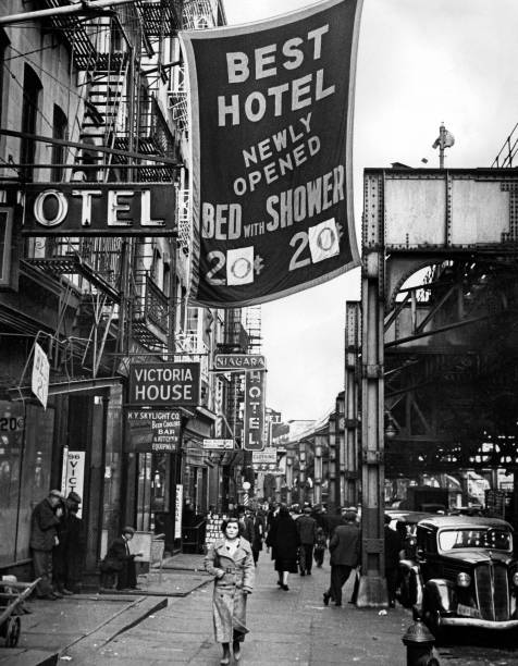 Several passers-by including a woman wearing a long coat stroll along the sidewalk in front of the hotels in front of the Bowery metro on January